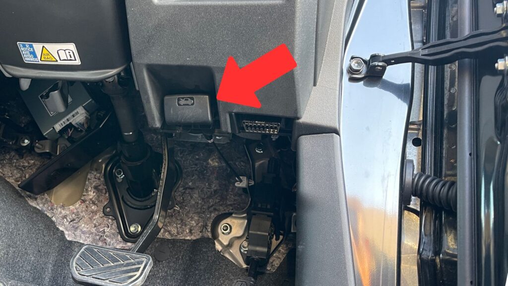 ジムニーJB64のボンネットオープナー位置。運転席足元、アクセルペダルの上にある黒いレバーを赤矢印で示した写真。