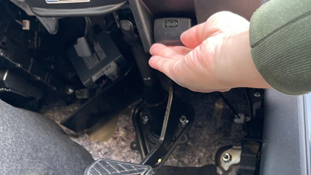 ジムニーJB64のボンネットオープナーレバーを手で引いている場面。運転席足元での操作の様子をわかりやすく撮影。