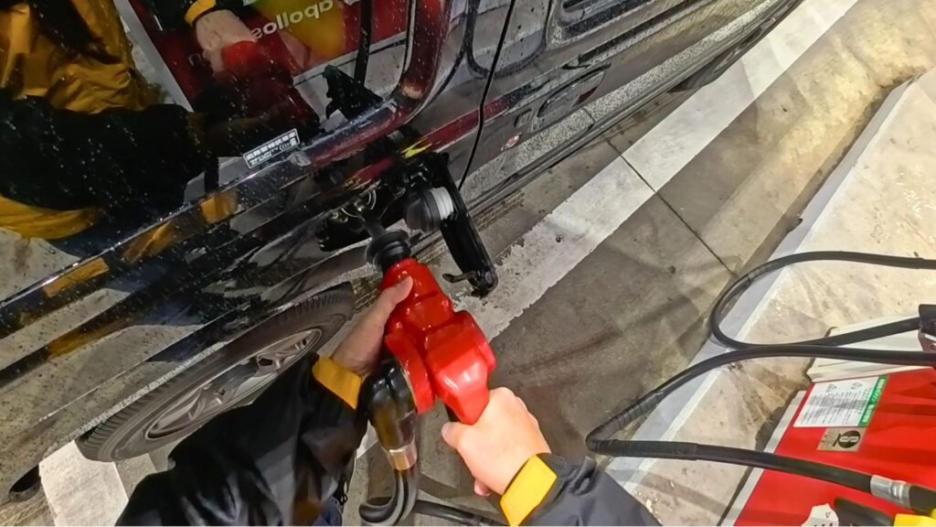 セルフガソリンスタンドにて、40年以上無事故のプロが「両手」で重い給油ノズルを保持し、SUV（ジムニーJB64）の給油口へ確実に差し込む手順を実演している一人称視点（POV）画像。筆者自身の頭部にマウントしたアクションカメラInsta360 Ace Pro 2で撮影。