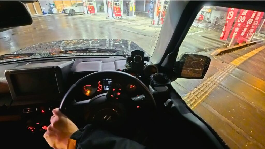 夜の雨の中、ガソリンスタンドの狭い給油レーンへ進入する、緊張感のある一人称視点（POV）画像。スズキ・ジムニーJB64の運転席から、筆者の頭部にセットしたアクションカメラInsta360 Ace Pro 2で撮影。セルフ給油が「怖い」と感じる初心者の心理的プレッシャーを表現している。