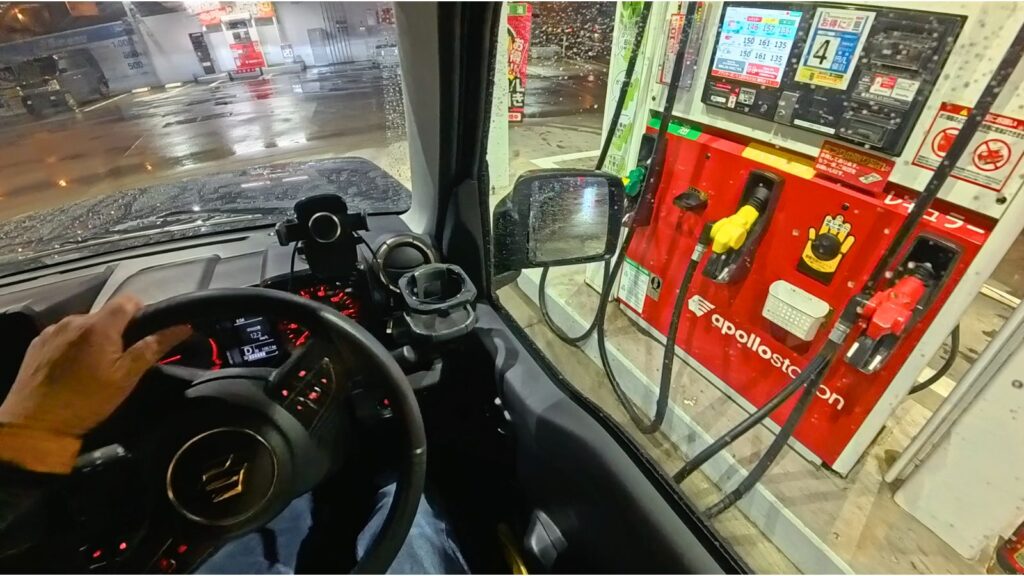 夜の雨のガソリンスタンドにて、スズキ・ジムニーJB64の運転席から、給油機との間に「約1m」の余裕を持って停車し、窓越しにノズルの色（油種）を確認している一人称視点（POV）画像。40年以上無事故のプロが推奨する「スマートな車寄せ」を筆者自身の頭部にマウントしたInsta360 Ace Pro 2で撮影。