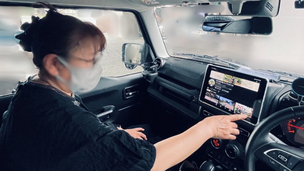 ジムニーJB64型の助手席に座る女性同乗者が、2DINスペースにマグネット固定されたiPad第9世代の画面を指で操作する様子。フロントガラスの背景は白くぼかされています。