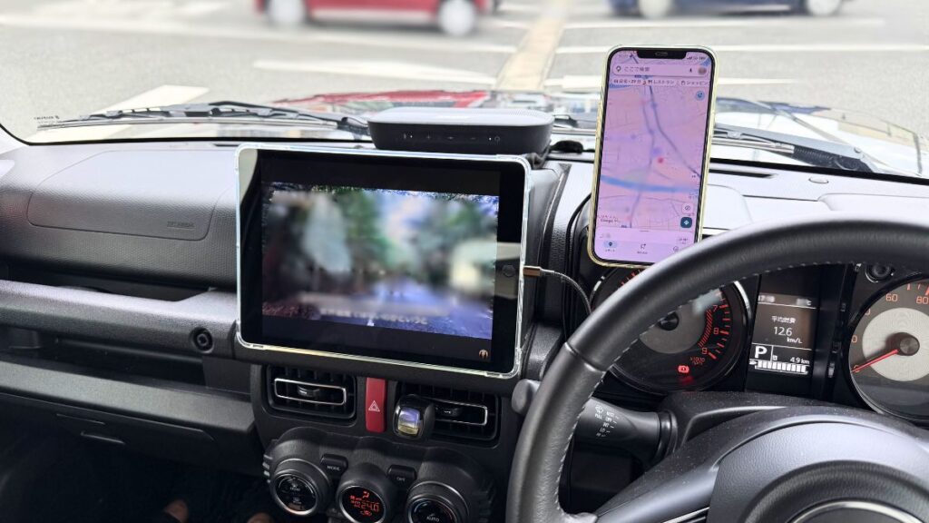 ジムニーJB64型の運転席メーターパネル、タコメーターのフード上部に装着された粘着吸盤式マグネットホルダーに、目的地設定画面を表示させたiPhoneを縦向きに固定した状態。2DINスペースにはiPad第9世代が横向きに配置されています。